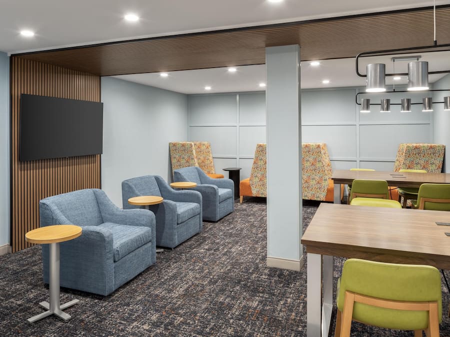 A modern hotel room with a flat-screen TV, a wooden panel wall, a seating area with sofas and round tables, and a conference table with chairs.