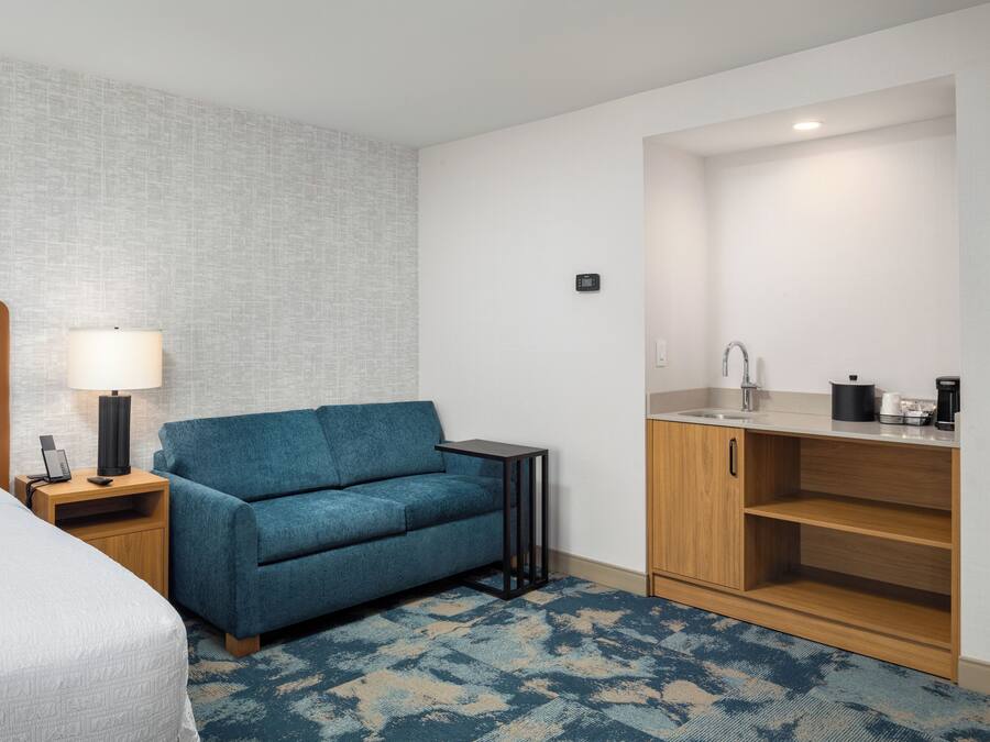A hotel room with a bed, a blue sofa, a small table with a lamp, a kitchenette with a sink and cabinets, and a patterned carpet.