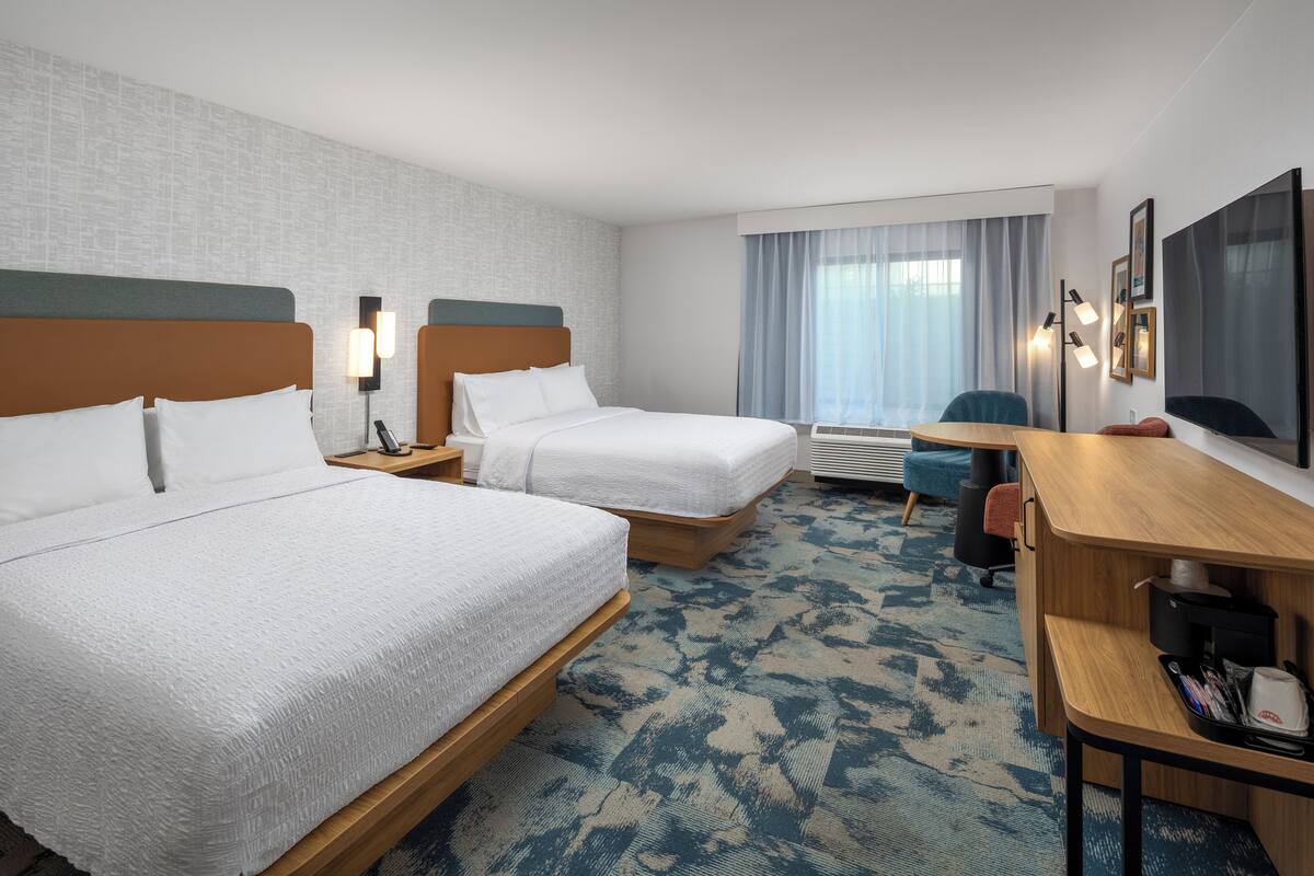 A hotel room with two beds, a desk, a chair, a flat-screen TV, and a large window with curtains.