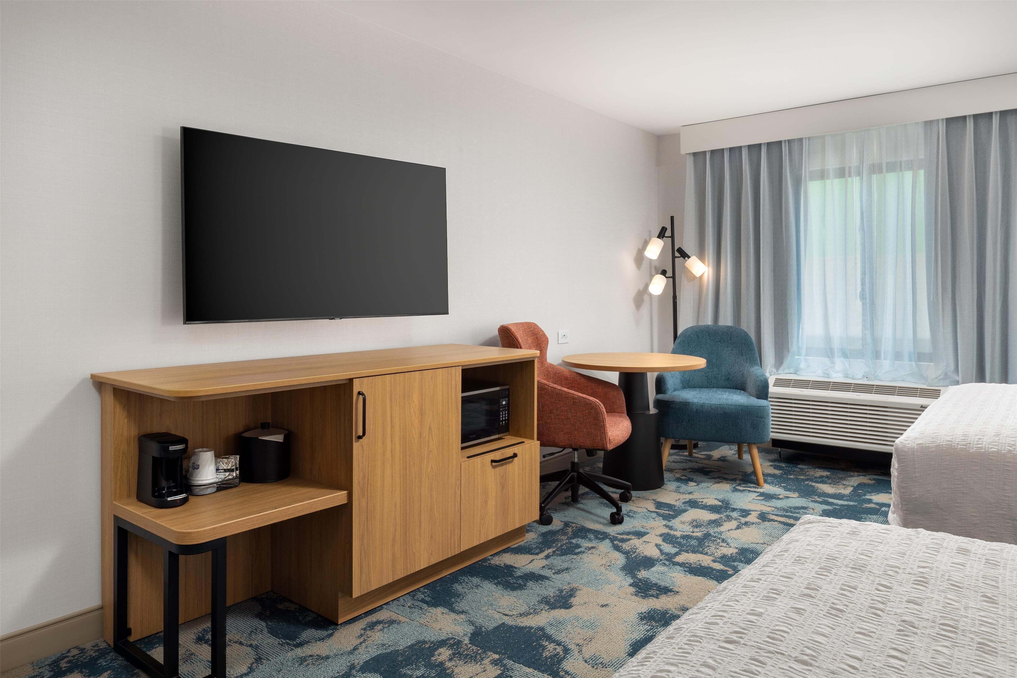Habitación de hotel con un escritorio de madera, una silla, una pequeña mesa redonda, una cama y un ventanal con cortinas.