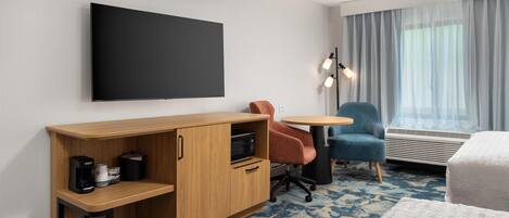 Habitación de hotel con un escritorio de madera, una silla, una pequeña mesa redonda, una cama y un ventanal con cortinas.