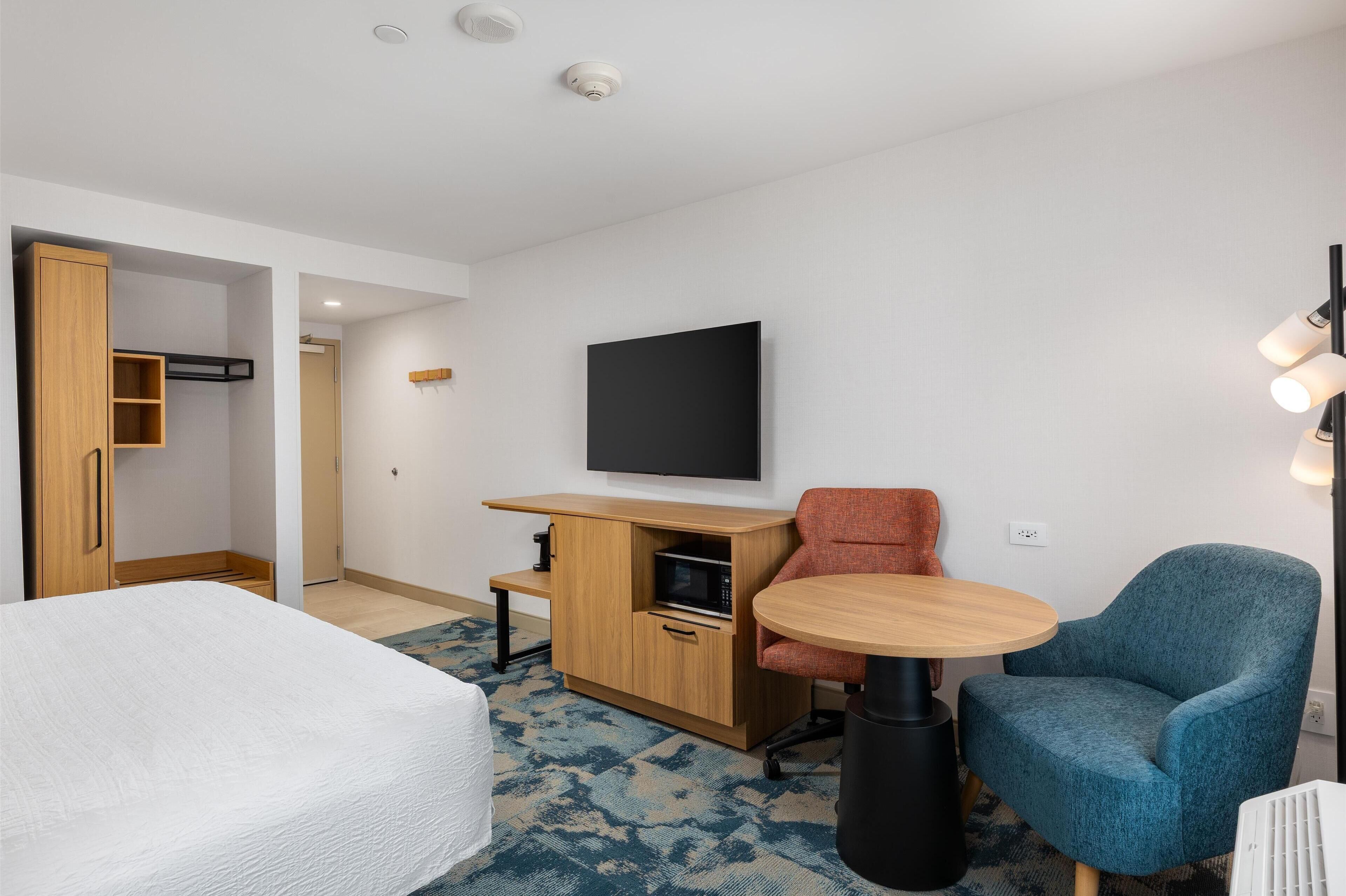 A compact hotel room with a bed, a small desk with a microwave, a TV, and two chairs.