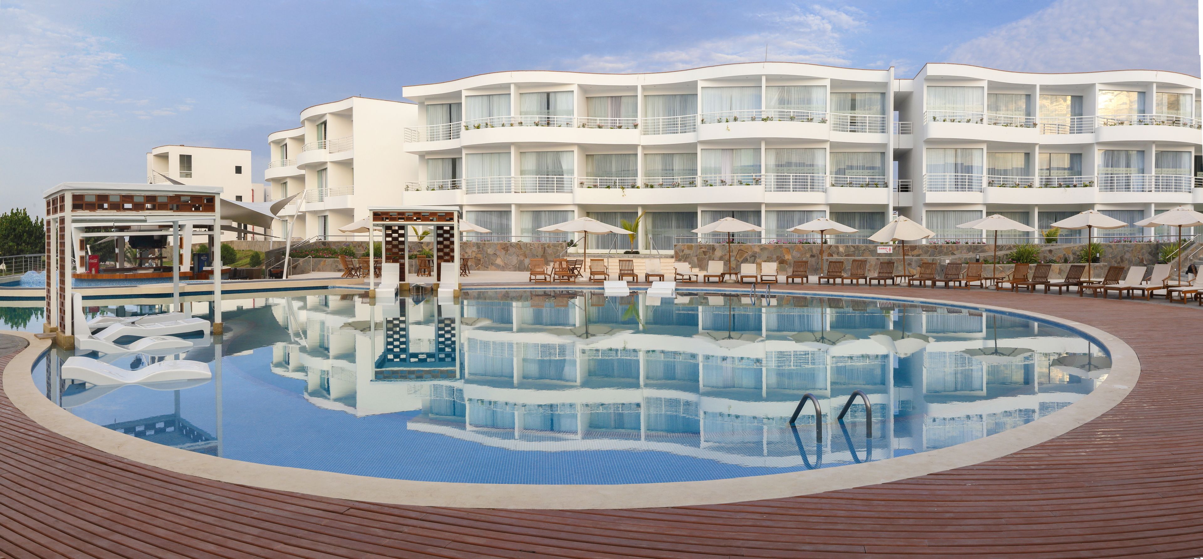 Una gran piscina con sillas de descanso y sombrillas, rodeada por un edificio hotelero de varios pisos con balcones.
