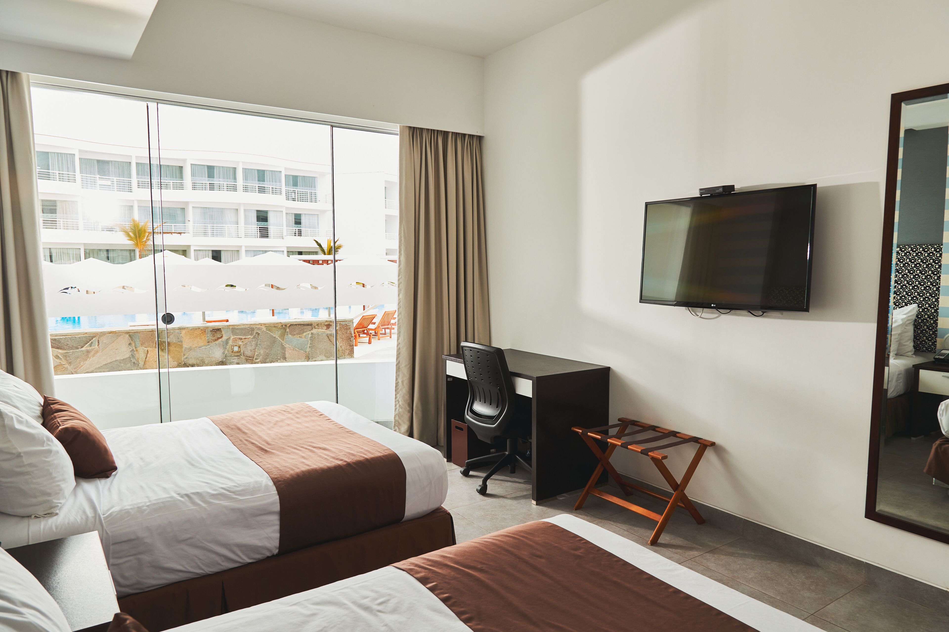 Une chambre d’hôtel avec deux lits, un bureau, une chaise, une télévision et une grande fenêtre donnant sur une piscine et des immeubles.