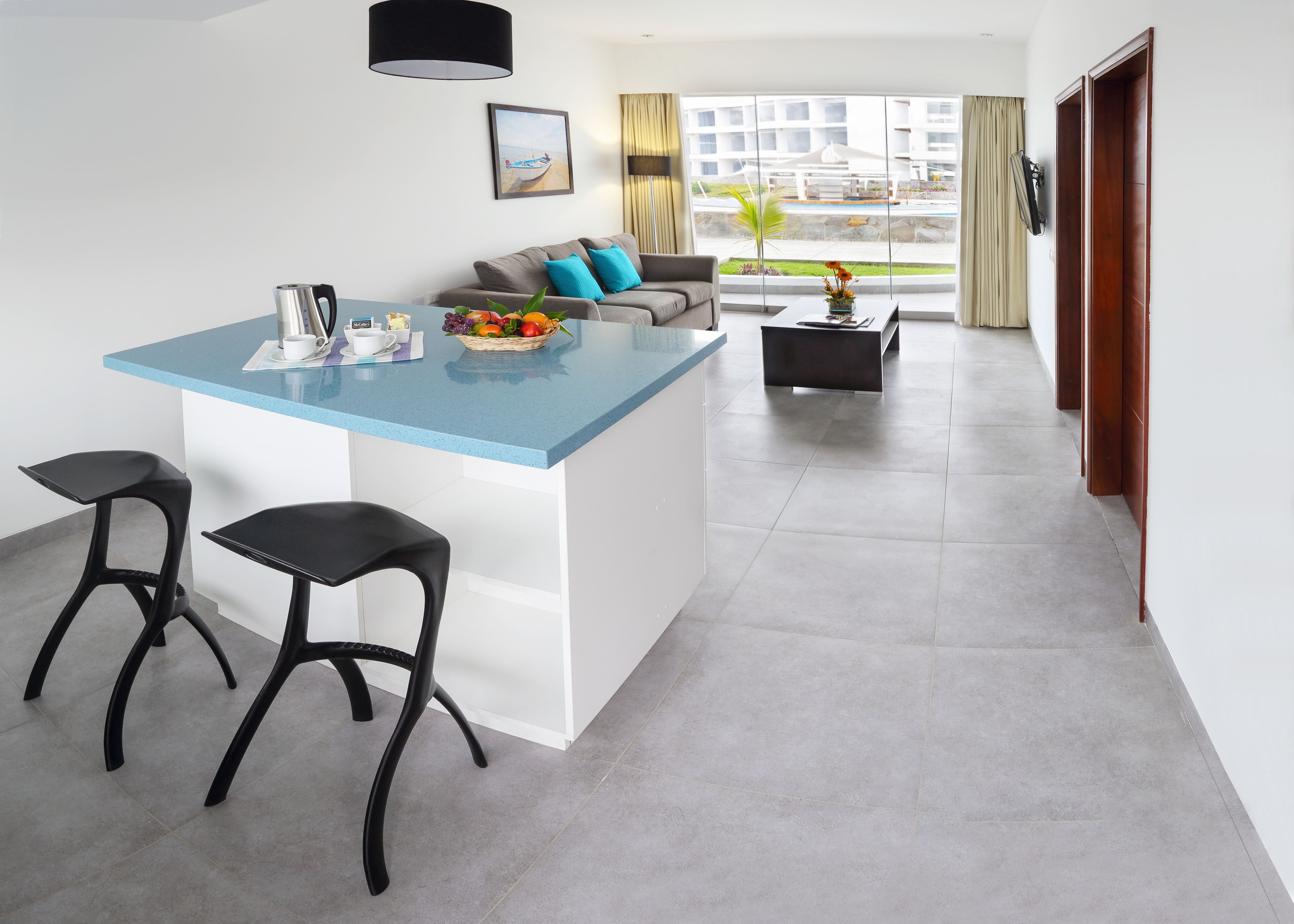 Una cocina moderna con encimera azul, dos taburetes negros, un sofá gris con cojines azules, y vistas a una piscina y edificios a través de amplias ventanas.