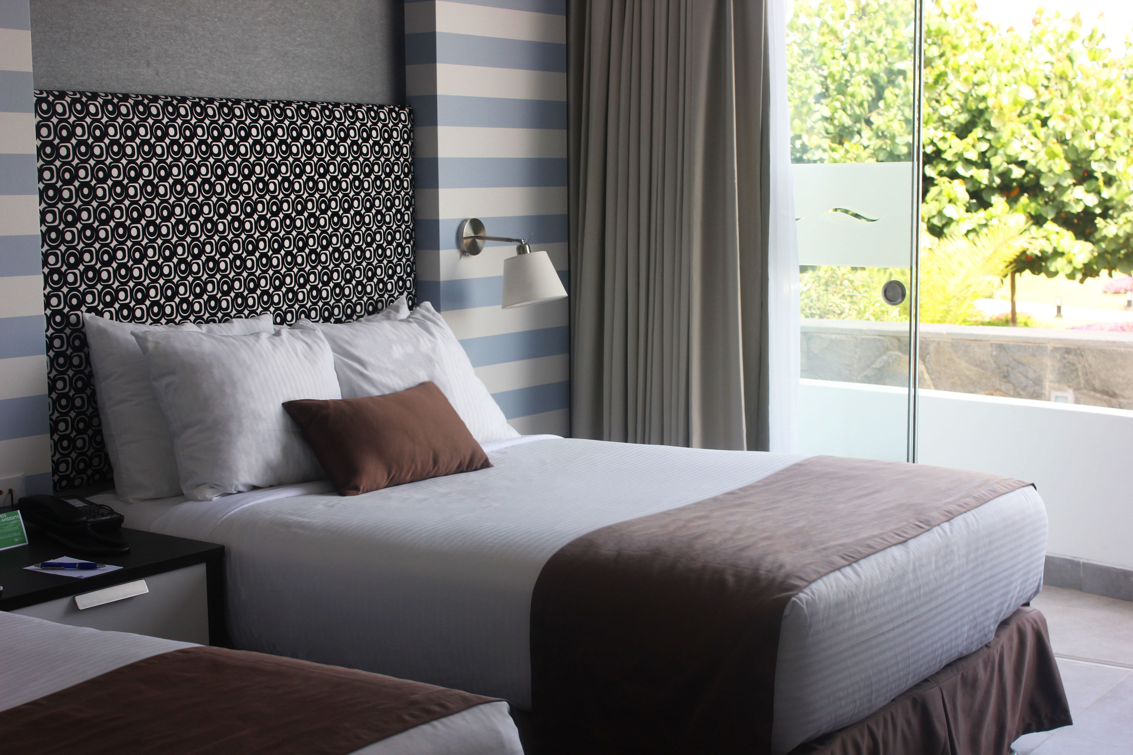 A hotel room with a large bed, a bedside table with a lamp, and a view of greenery outside.