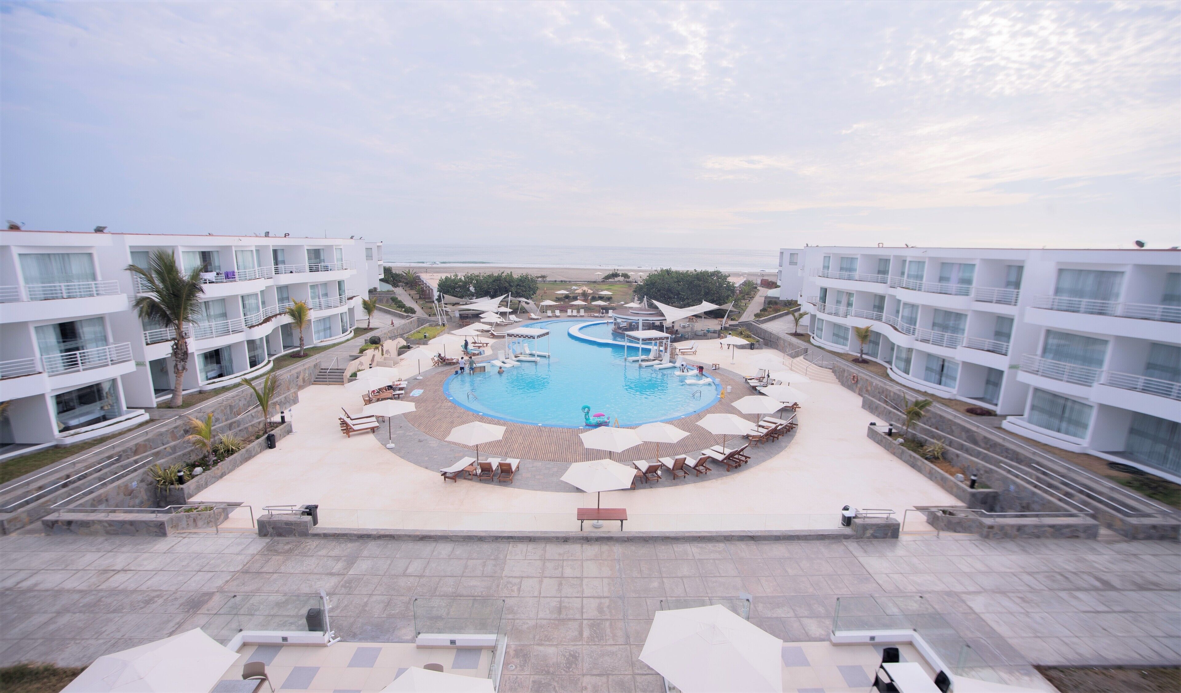 Un resort con una piscina central rodeada de sillas de descanso y sombrillas, edificios modernos con balcones y vista a la playa.