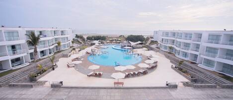 Un resort con una piscina central rodeada de sillas de descanso y sombrillas, edificios modernos con balcones y vista a la playa.