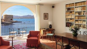 A room with a large window overlooking a body of water, a balcony with outdoor furniture, a bookshelf, and a dining area. - Hôtel La Ponche (Saint-Tropez)