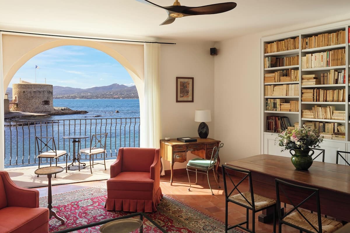Una habitación con un ventanal grande que da a una masa de agua, un balcón con mobiliario exterior, una estantería y un comedor.