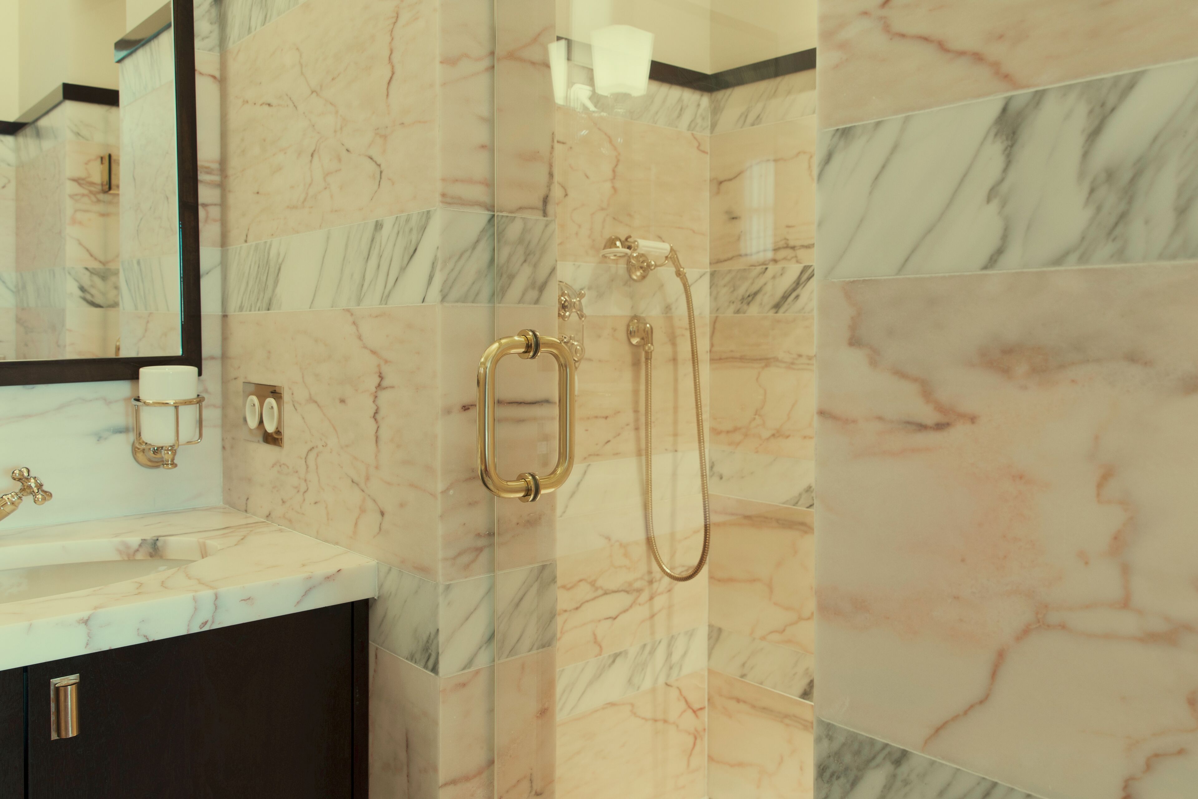 Une salle de bain carrelée de marbre, avec une pomme de douche dorée et un miroir.