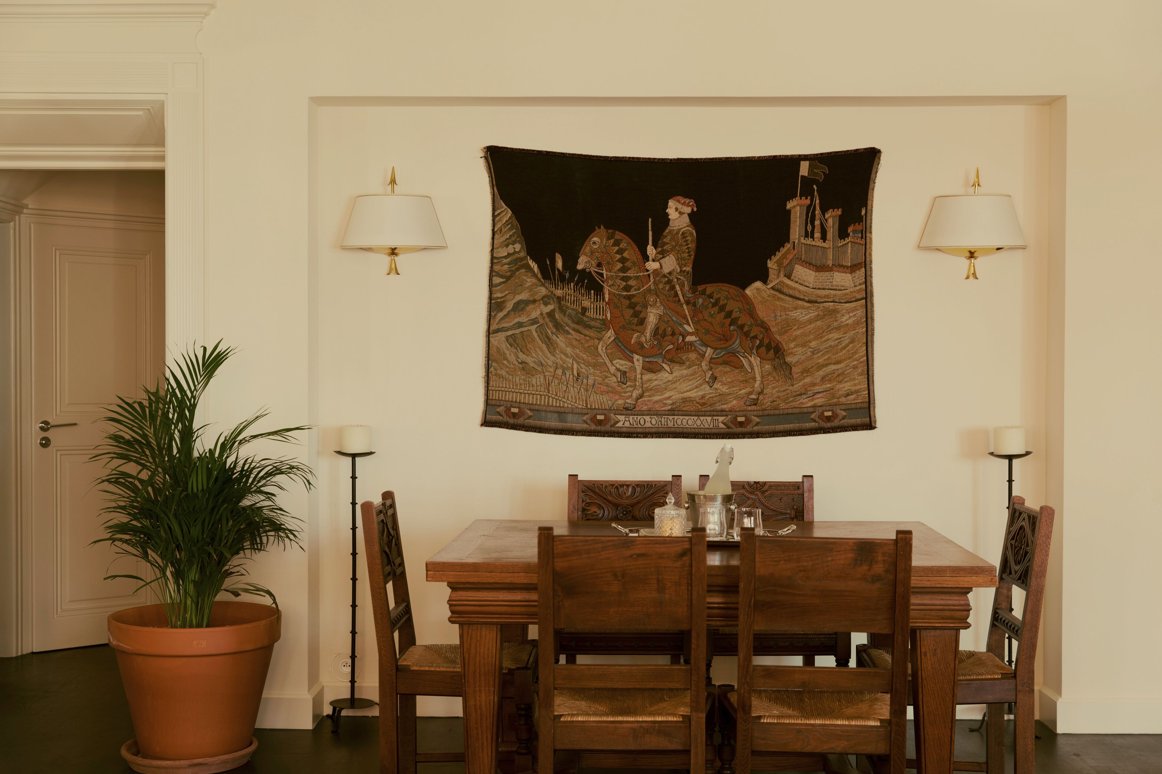 Uma área de refeições com uma mesa posta para quatro pessoas, cercada por cadeiras de madeira, um vaso de planta e uma tapeçaria na parede.