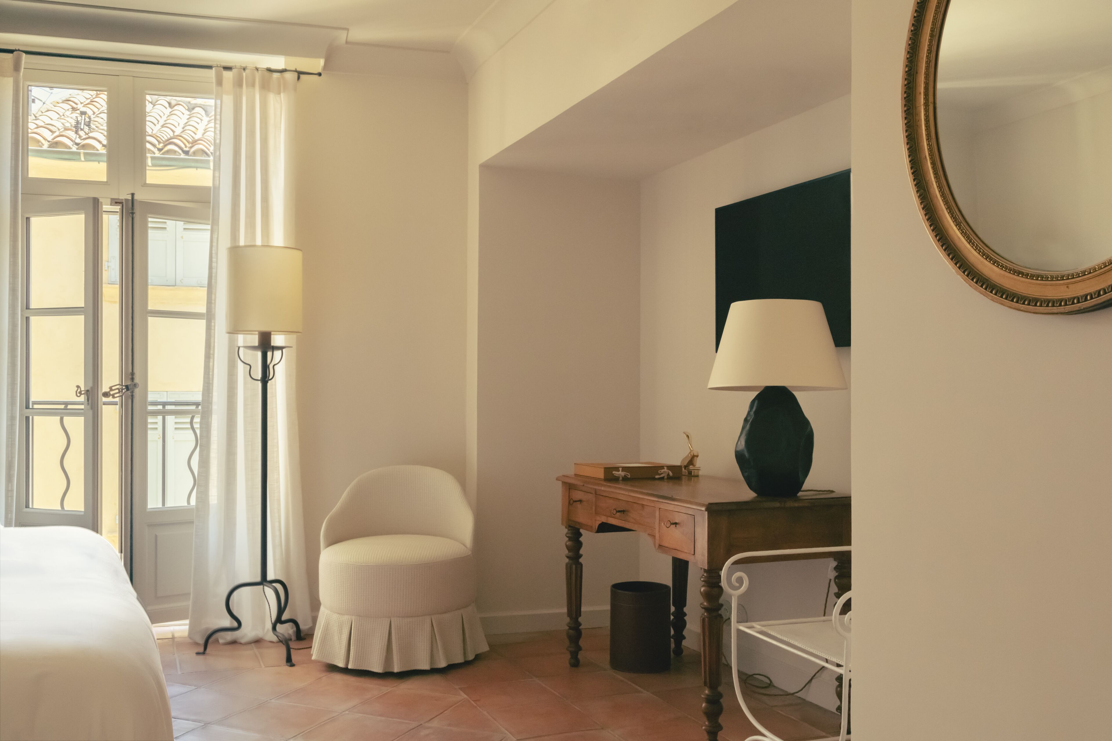 Una camera d'albergo con un letto, una sedia, una scrivania con una lampada, uno specchio e vista attraverso una finestra con tende.