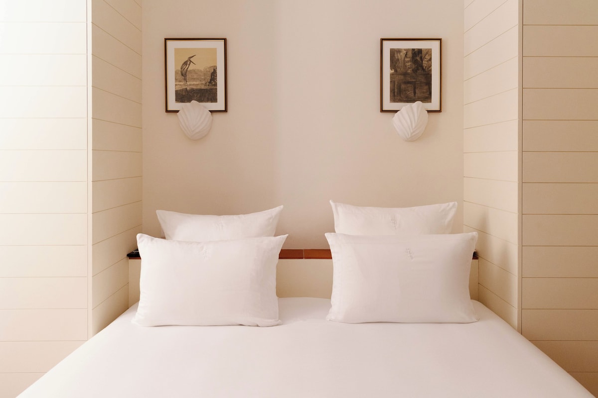 A neatly made bed with white bedding and pillows, two framed pictures on the wall, and a white shell decoration.