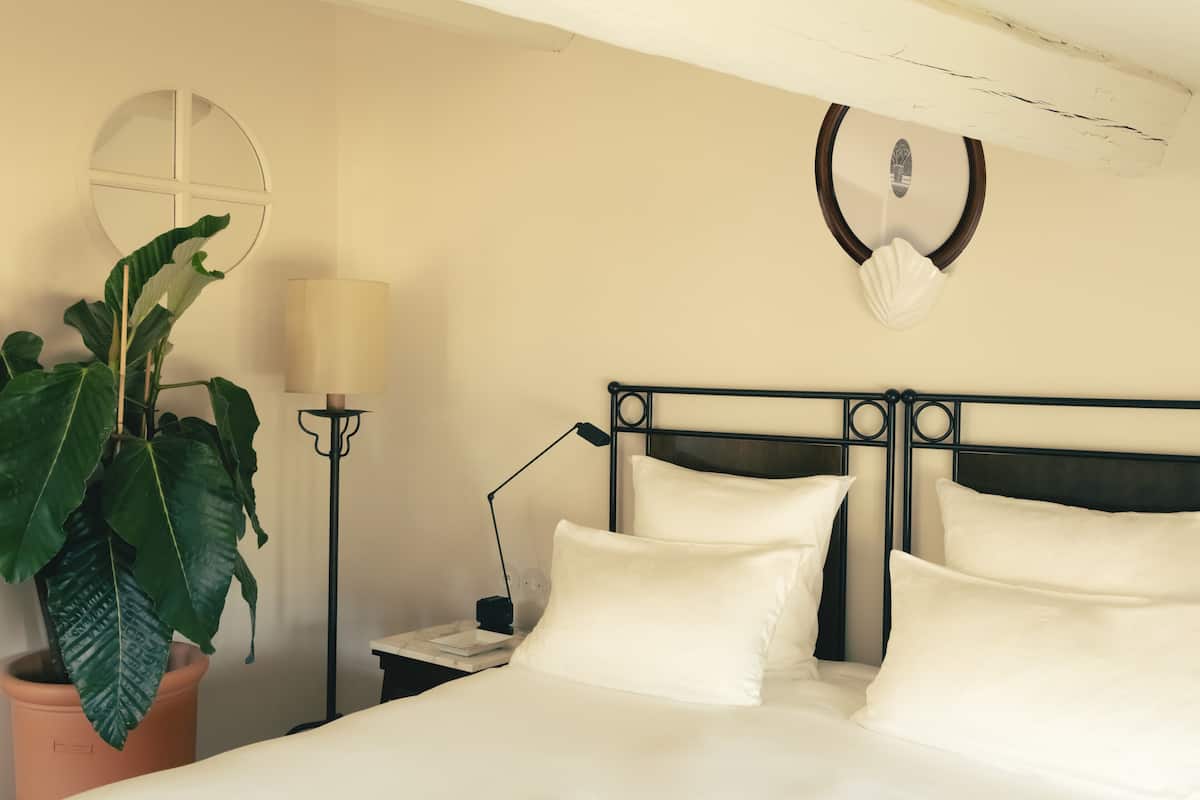 Una cama bien hecha con sábanas blancas, una planta en maceta, una lámpara de noche y un adorno en la pared.