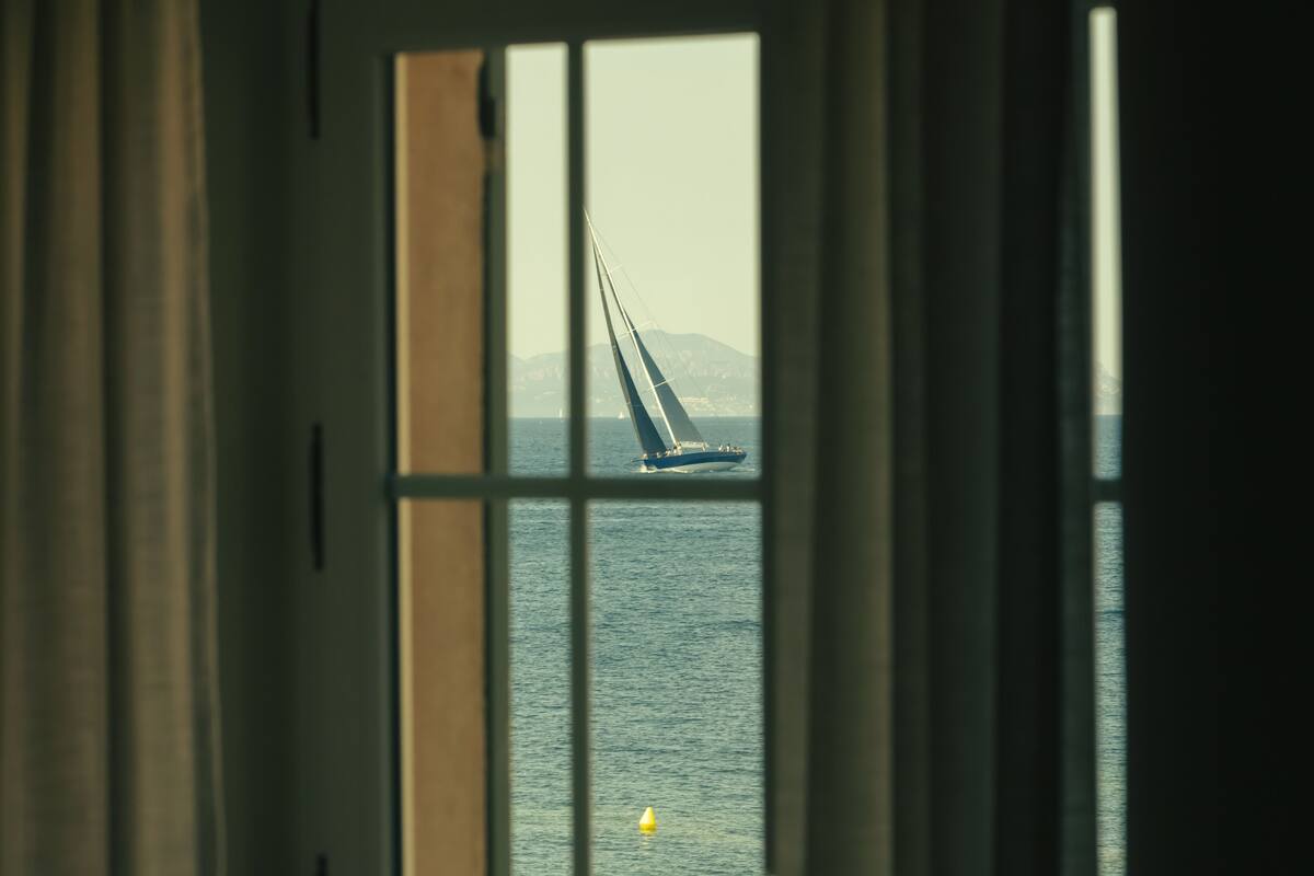 Un velero en el agua, visible a través de una ventana que ofrece vistas al mar y montañas distantes.