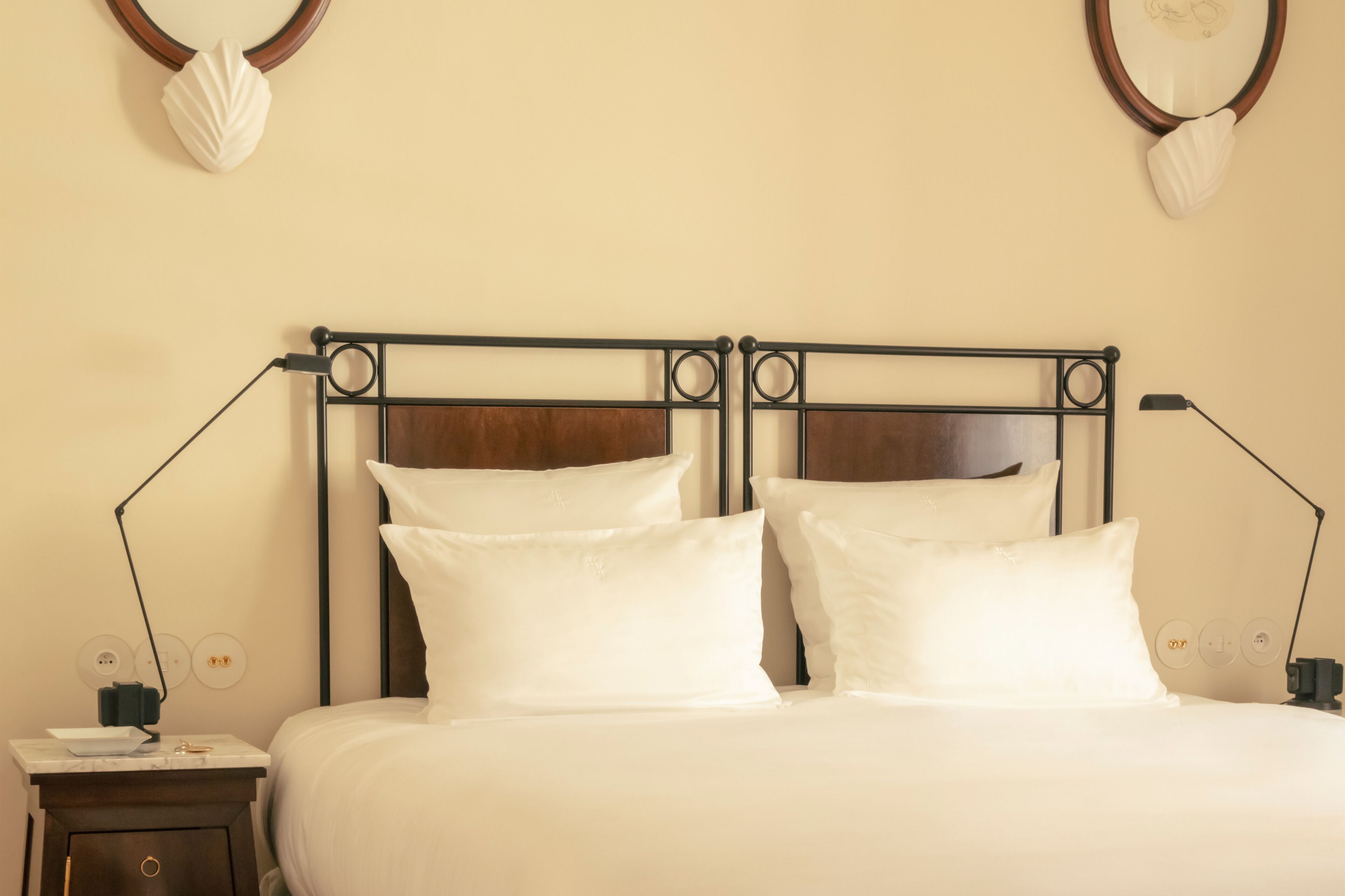 A neatly made bed with a metal headboard, two bedside lamps, and a small bedside table with a lamp and a power outlet.