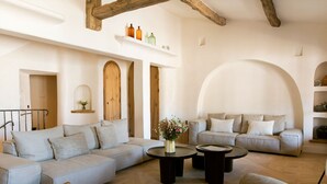 A spacious living room with a light grey sofa, a round coffee table, and a wooden chair. There are two sofas facing each other, a wooden beam ceiling, and a white wall with a shelf holding bottles. - Hôtel La Ponche (Saint-Tropez)