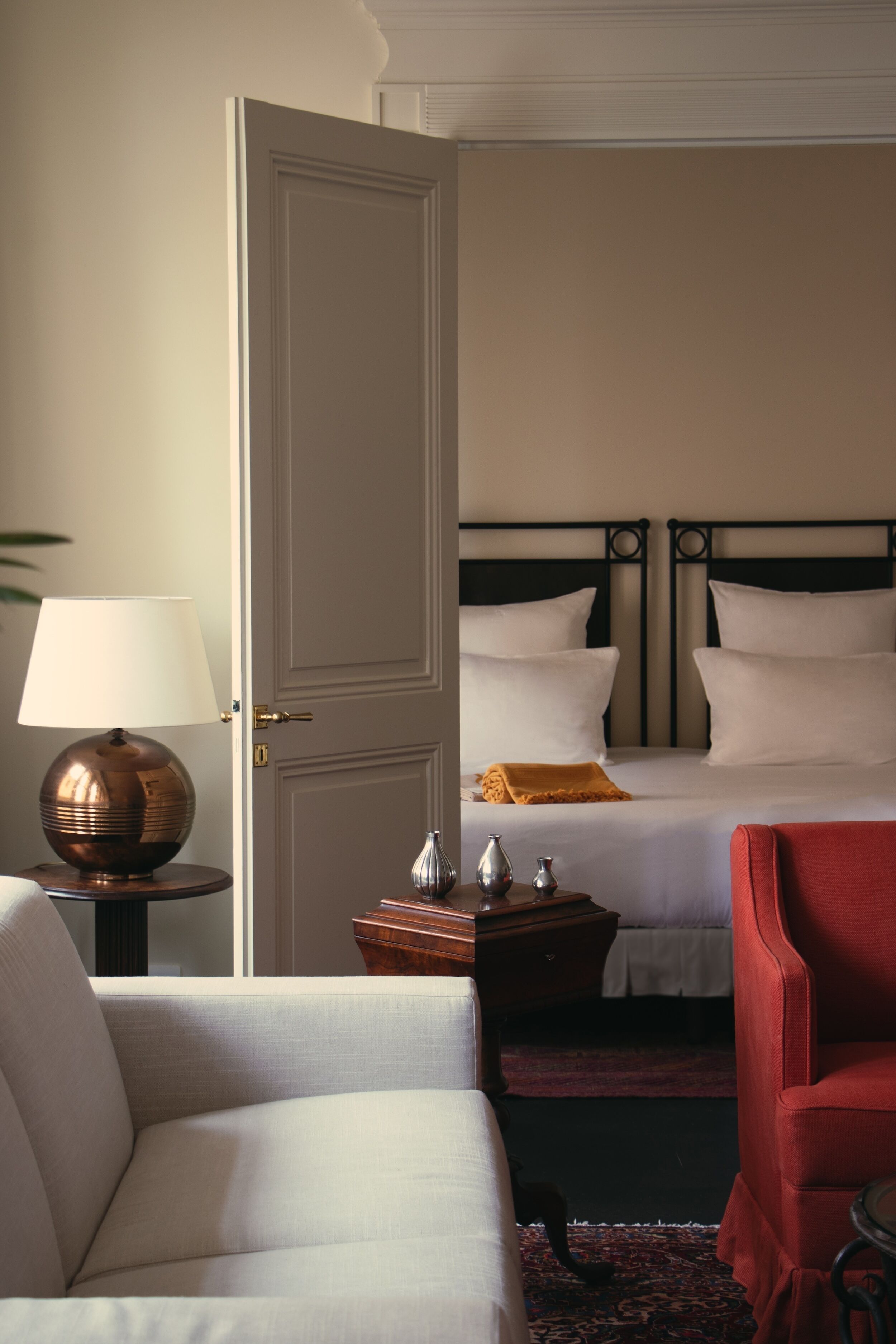 Quarto de hotel com duas camas, um espaço de estar com uma poltrona, uma luminária e uma mesinha de centro com objetos decorativos.