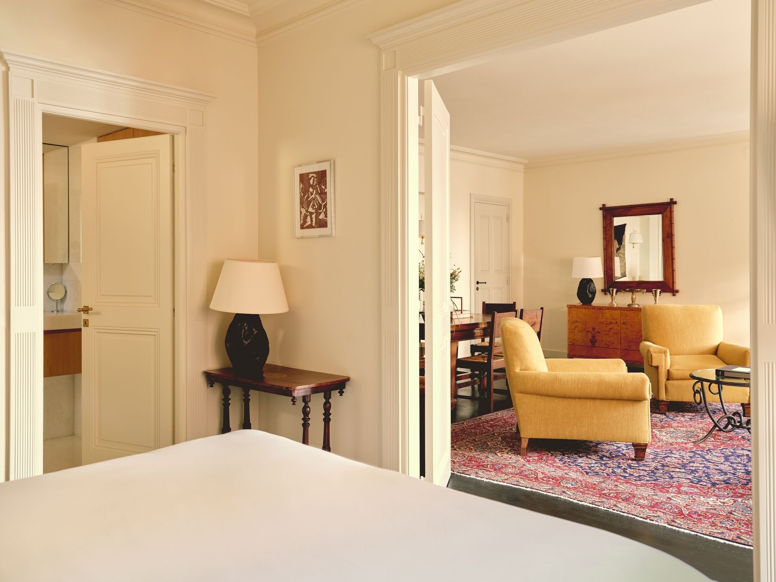 A hotel room with a bed, a desk with a lamp, two armchairs, a dining table with chairs, and a large mirror.