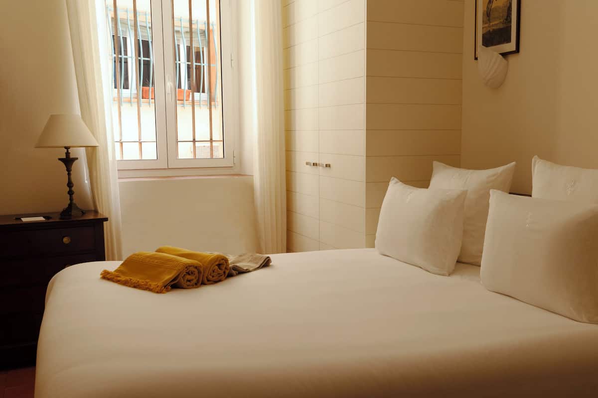 Una habitación de hotel con cama, mesita de noche, lámpara y ventana con cortinas.