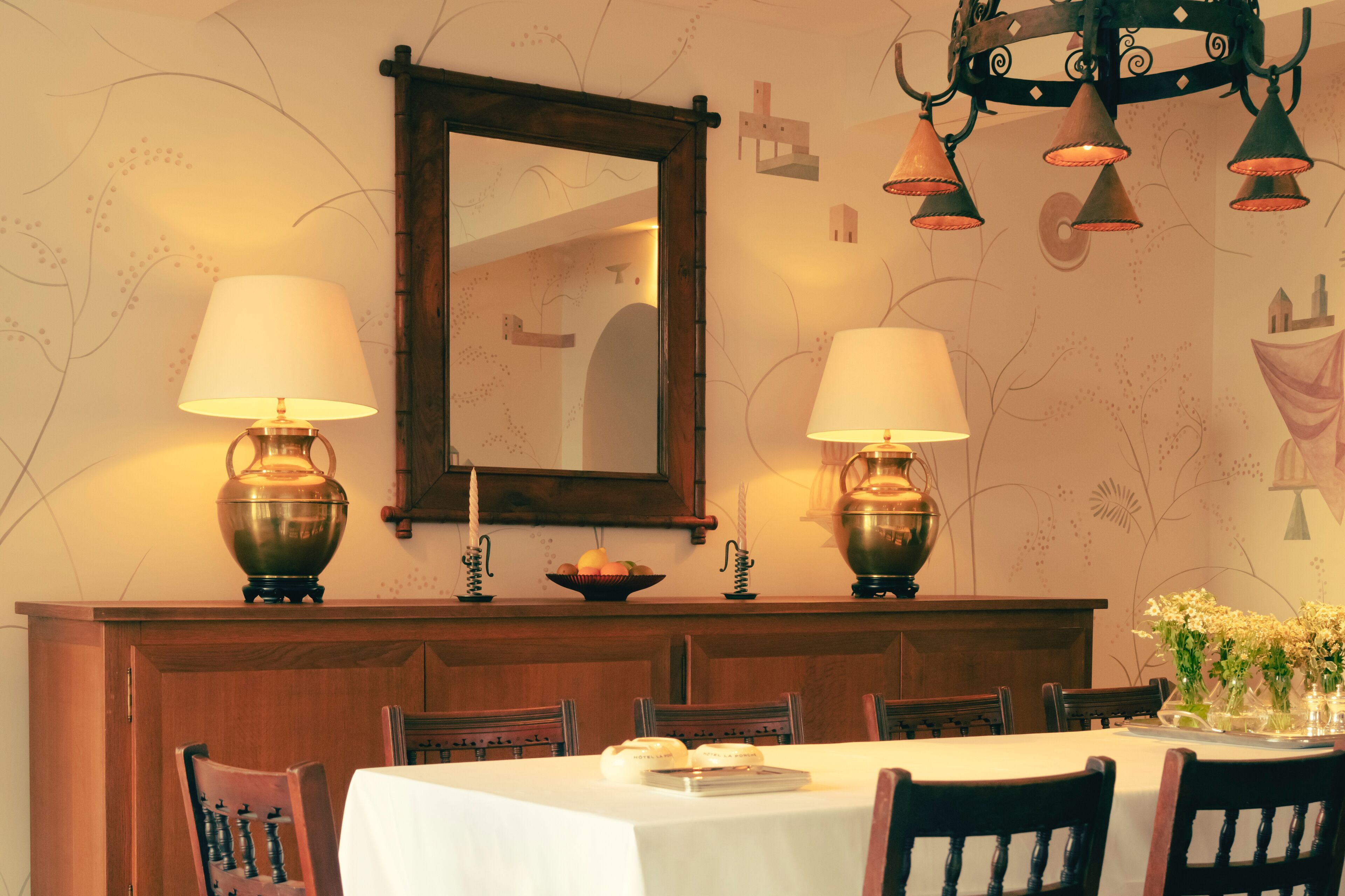 A dining area with a wooden table, chairs, and a mirror above. There are two lamps on the wall and a vase with flowers on the table.