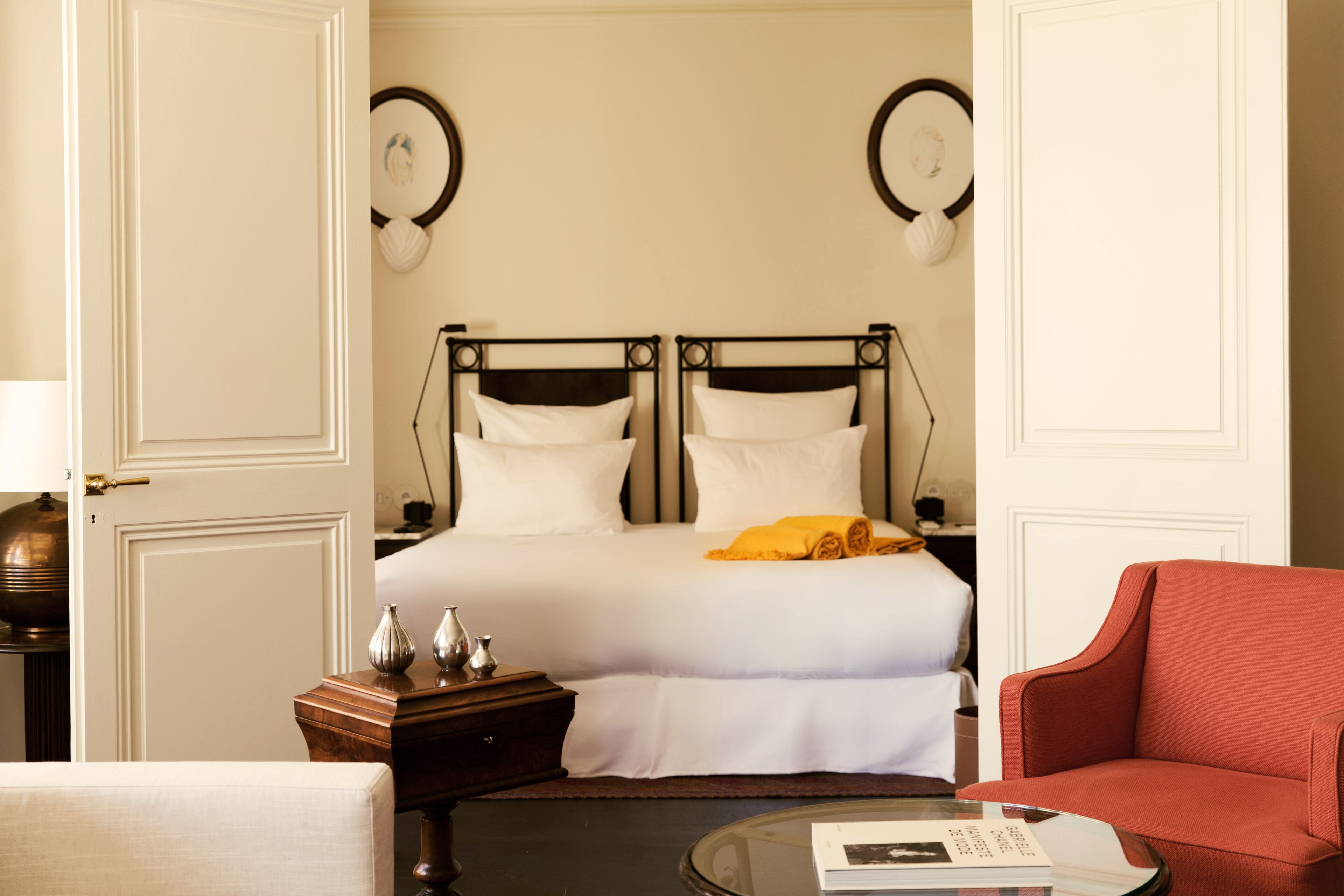 Una habitación de hotel con una cama, un sillón rojo, una mesa de centro de cristal y una mesita de noche con lámpara.
