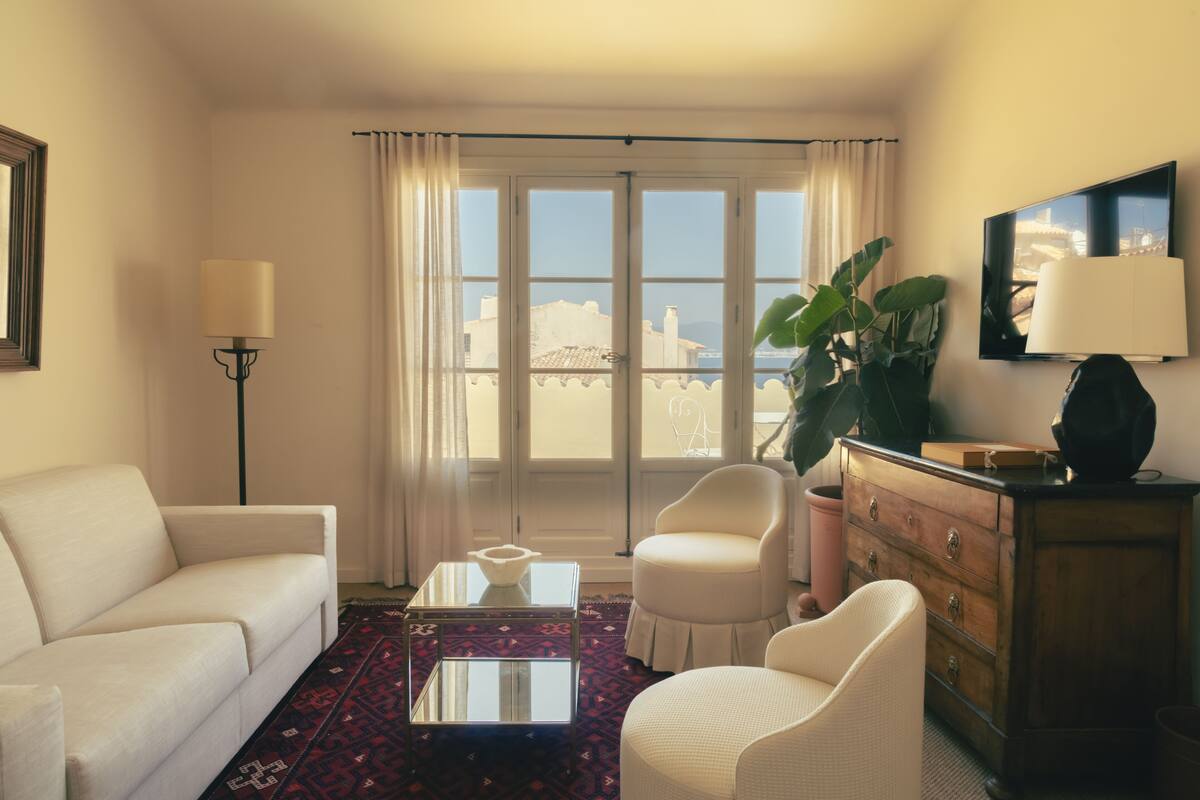 Habitación de hotel con sofá blanco, una mesita de centro de vidrio, cómoda de madera, una planta grande en maceta y vistas a través de un ventanal.