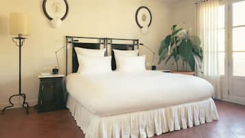 Ein großes Bett mit weißer Bettdecke und Rüschelvorderseite, ein Nachttisch mit Lampe, ein großer Topfpflanze und zwei Wanduhren.