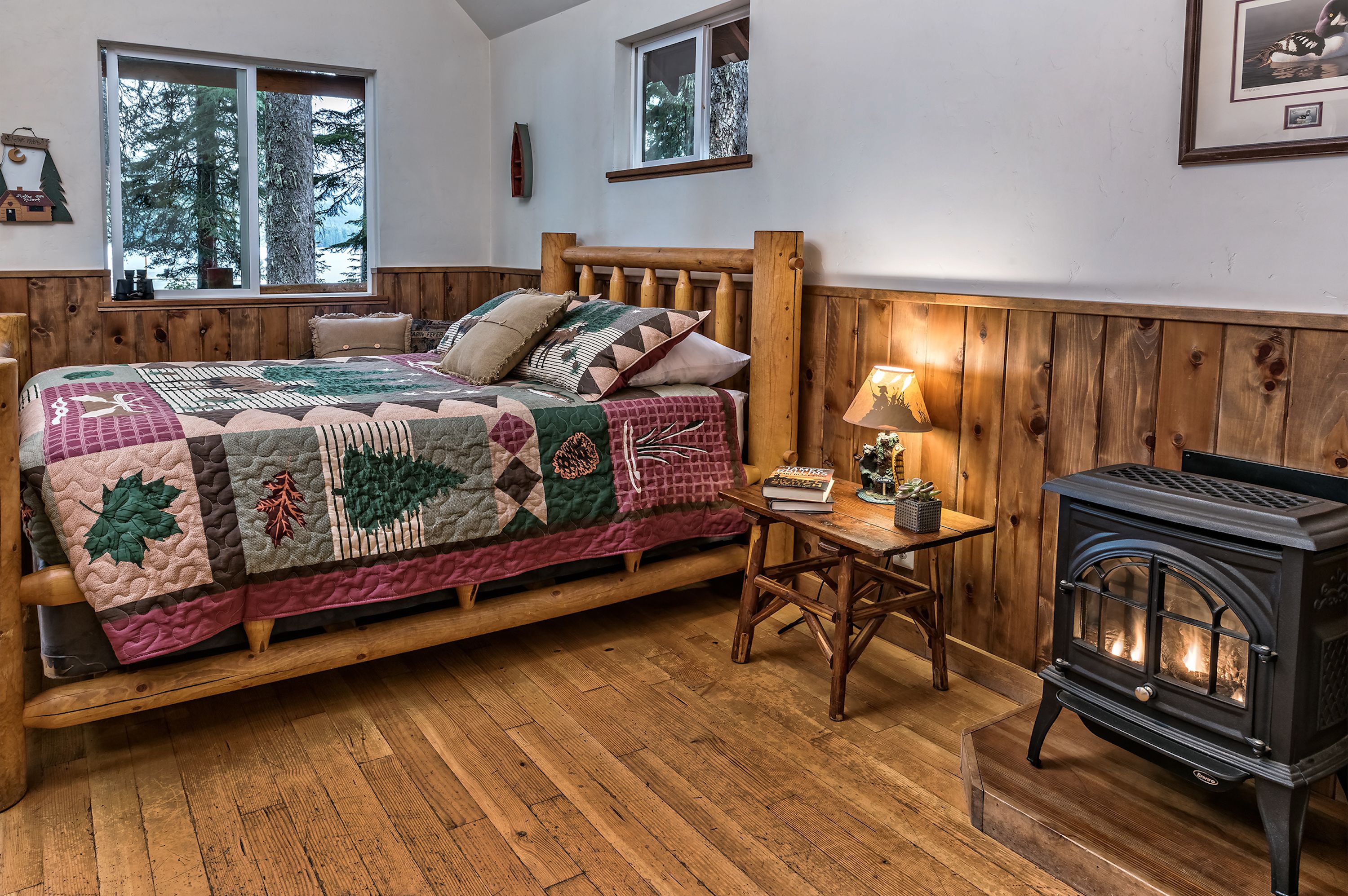 A cozy room with a wooden bed, a quilt, a small table with a lamp, and a wood-burning stove.