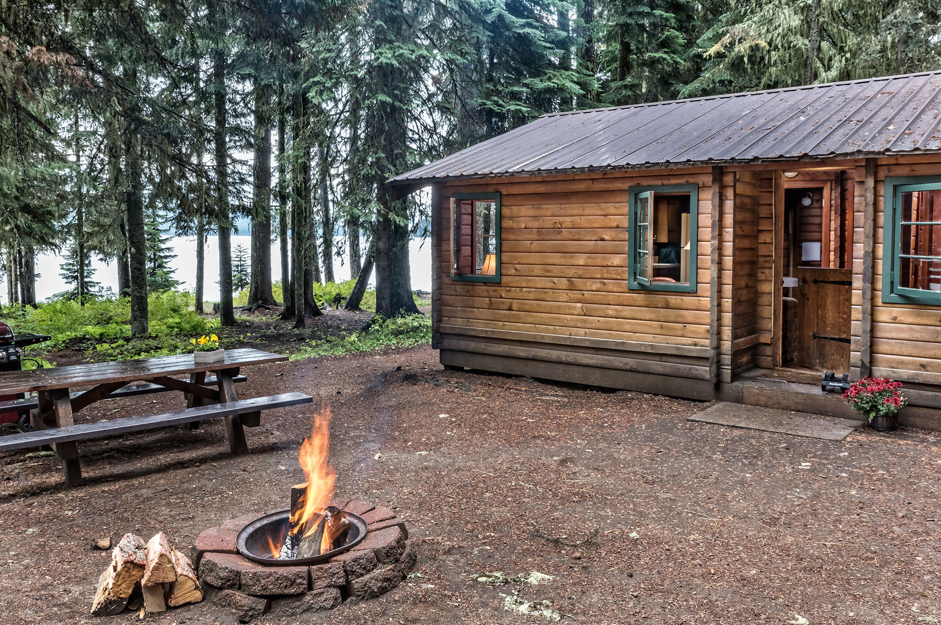 A rustic cabin with a fire pit, wooden deck, and a view of a forested lake.