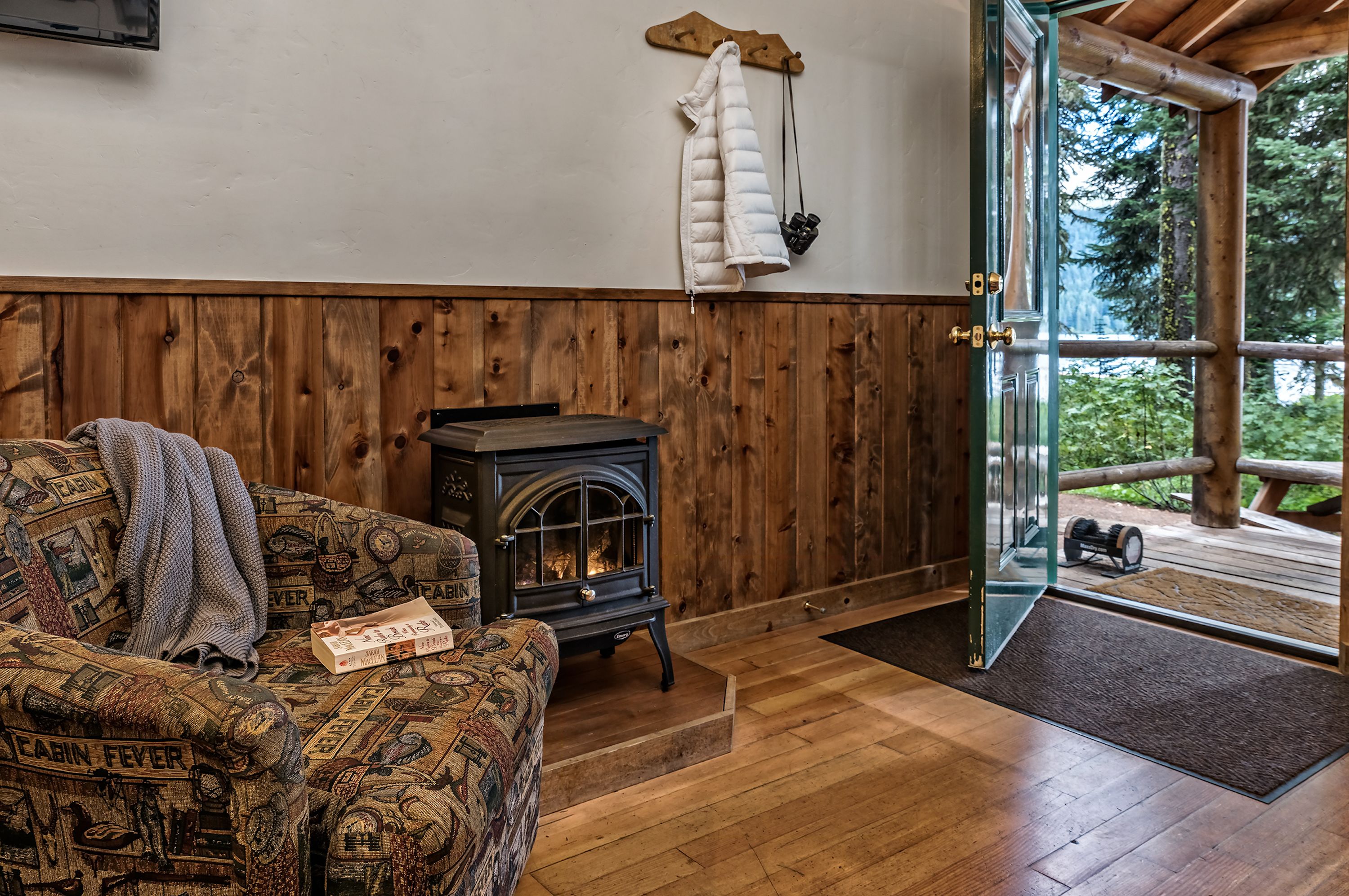 Interior de una cabaña con un sofá estampado, un estufa de leña y una puerta que da a una terraza exterior.