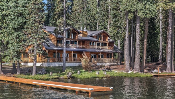 Una cabaña grande de madera con varios balcones, ubicada junto a un lago rodeado de altos árboles.
