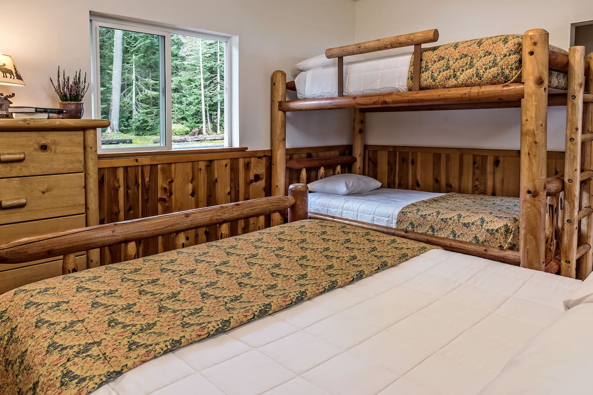 Een stapelbed met houten frame, een dressoir met spiegel en een raam met uitzicht op bomen.