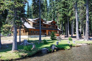 Un chalet doté d’une grande terrasse, entouré de grands arbres et traversé par un petit ruisseau.