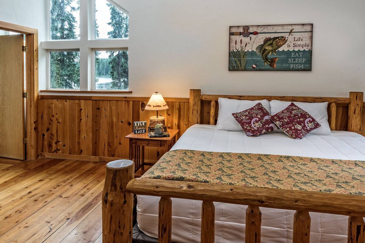 Een slaapkamer met houten details, een bed, nachtkastje, lamp en een raam met uitzicht op bomen.
