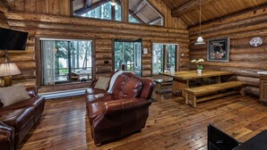 Interior de una cabaña de troncos con suelo de madera, sofá de cuero, mesa de comedor y grandes ventanales con vistas al bosque.