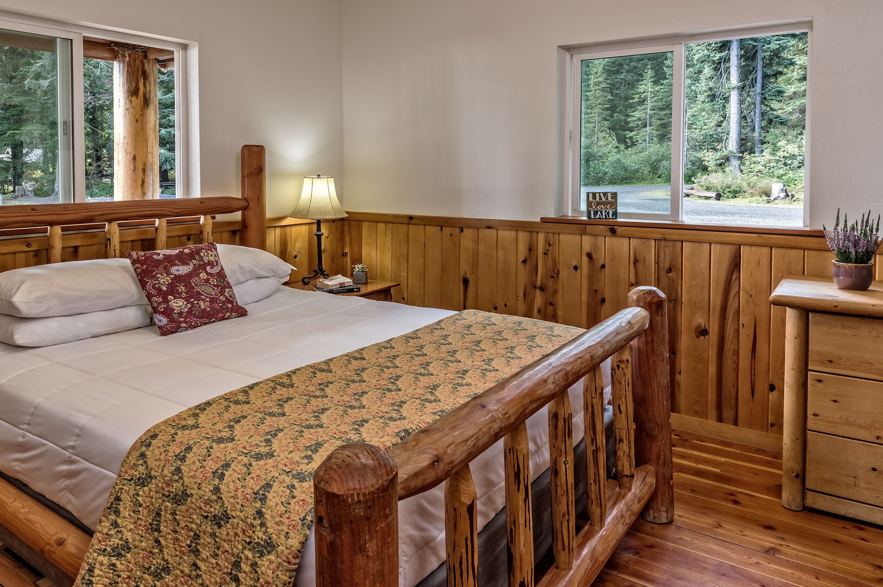 Una cama de madera con una colcha estampada con motivos florales, una mesita de noche con una lámpara, y vistas a un bosque a través de amplios ventanales.