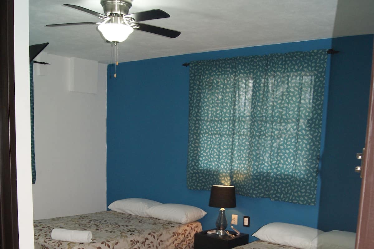 A room with two beds, a ceiling fan, a nightstand with a lamp, and a window with patterned curtains.