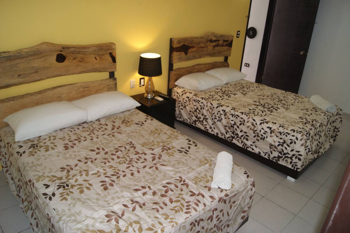 A hotel room with two beds, each with a floral bedspread, a wooden headboard, a nightstand with a lamp, and a towel on the floor.