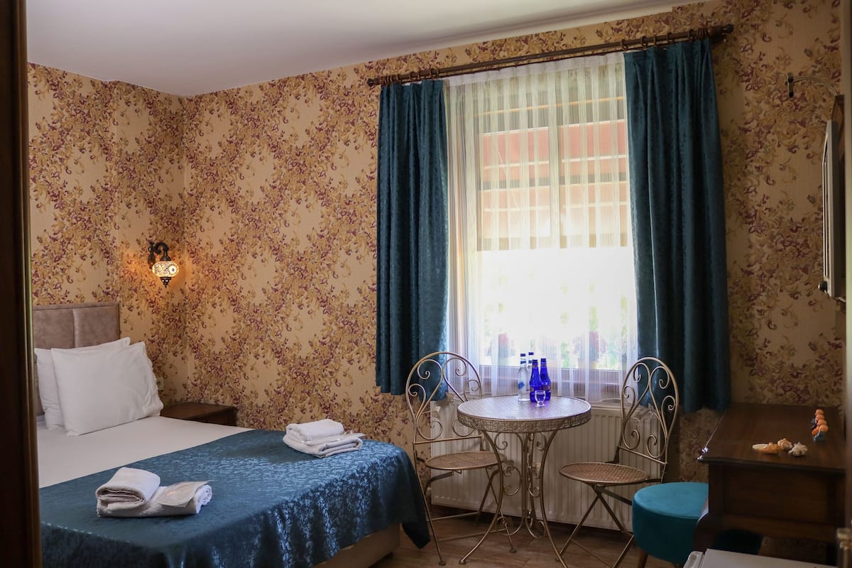 Quarto de hotel com cama, mesa redonda e cadeiras, pequena escrivaninha e janela com cortinas.