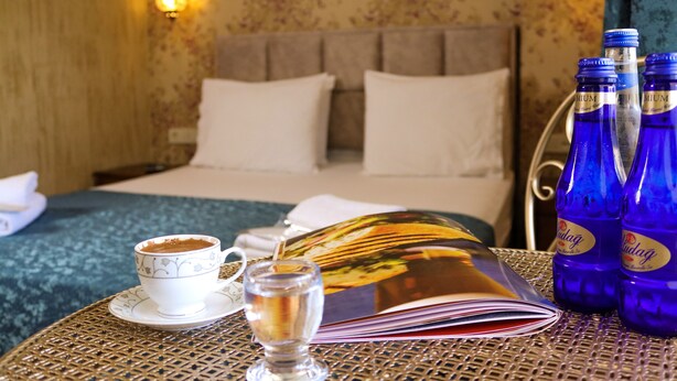 A neatly made bed with white linens, a table with a book, a cup of coffee, and two bottles of water. - Doruk Hotel (Ayvalik)