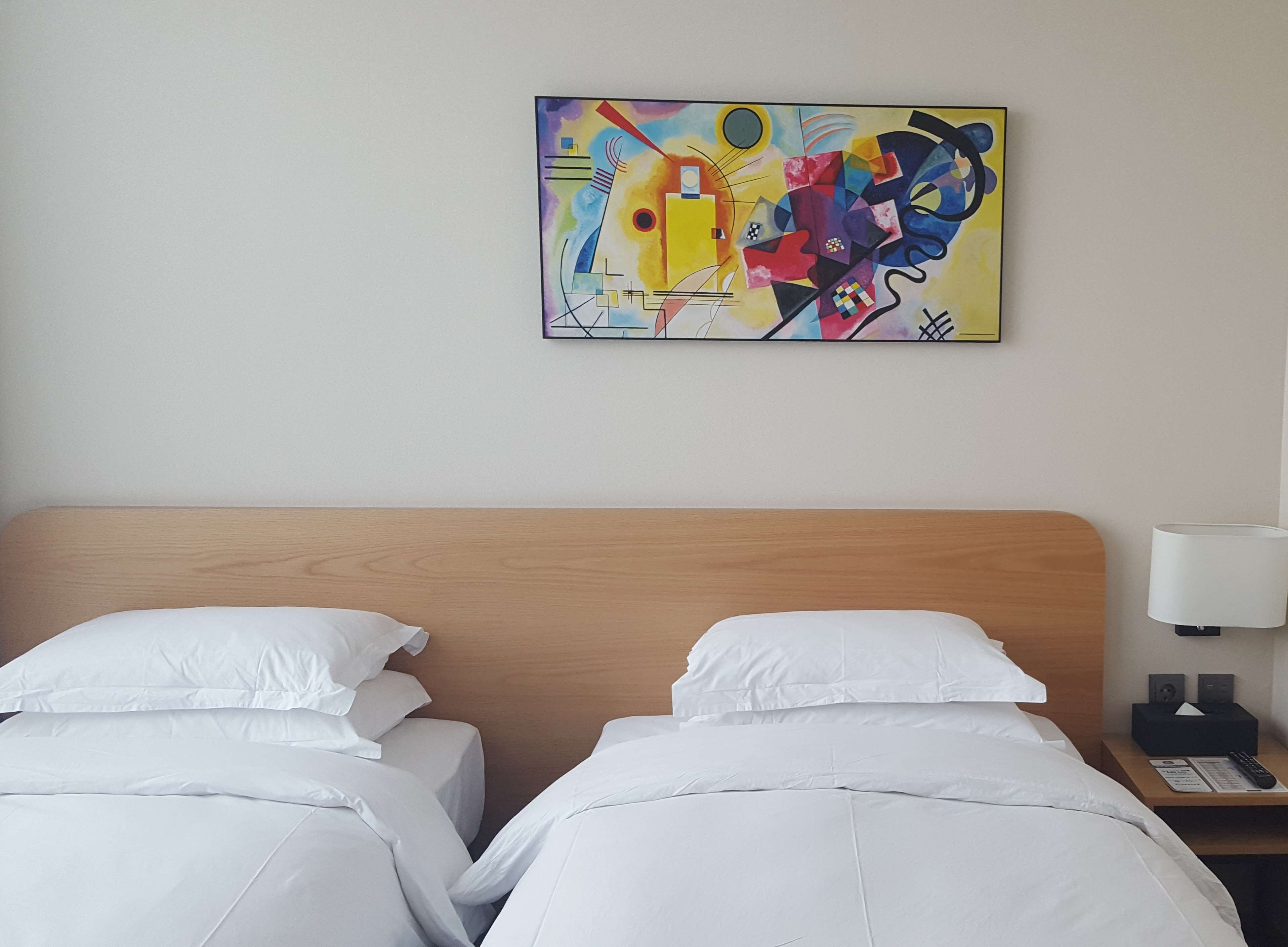 A hotel room with two beds, a wooden headboard, a nightstand with a lamp, and a colorful abstract painting on the wall.