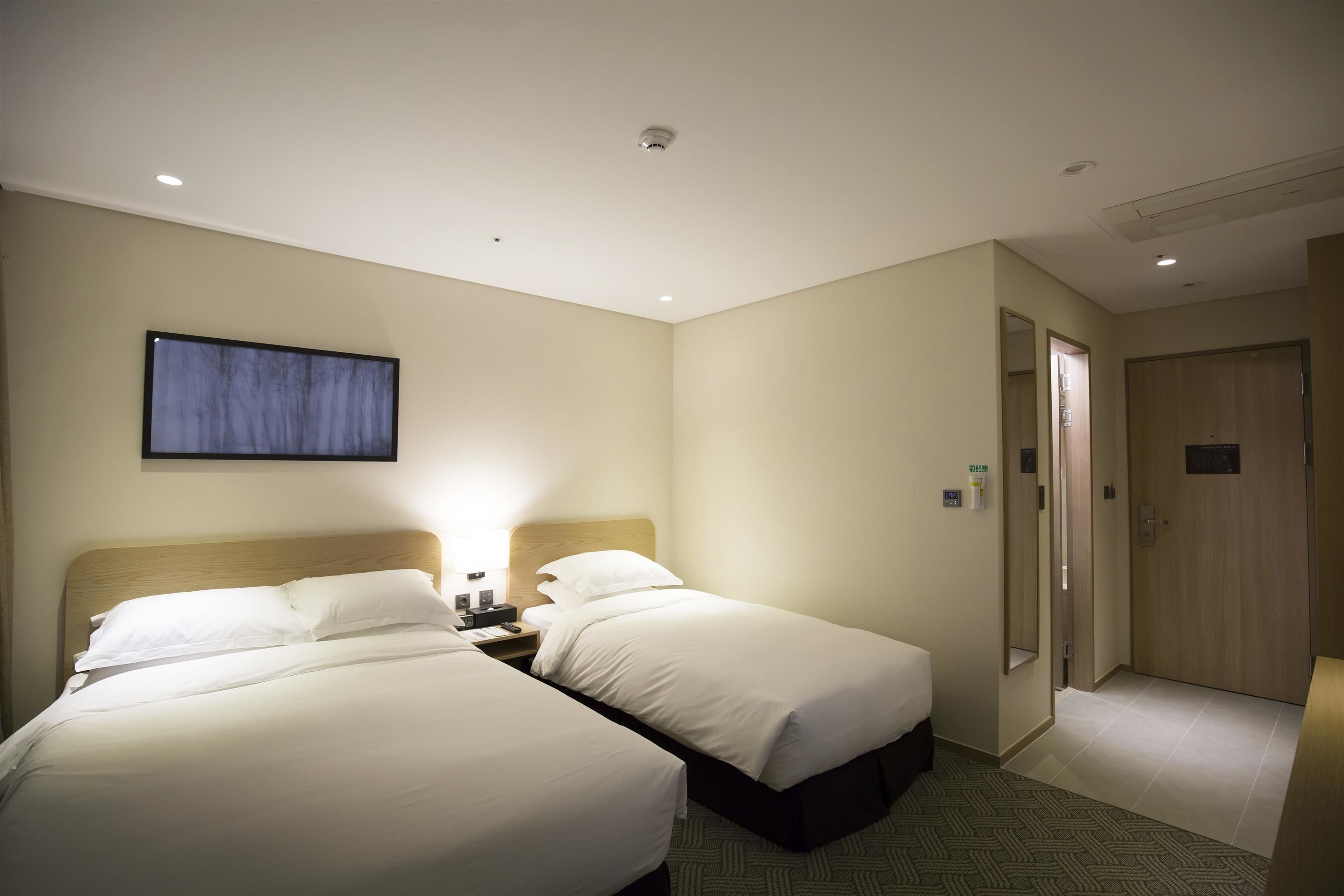 A hotel room with two beds, a nightstand, a wall-mounted lamp, and a door leading to another room.