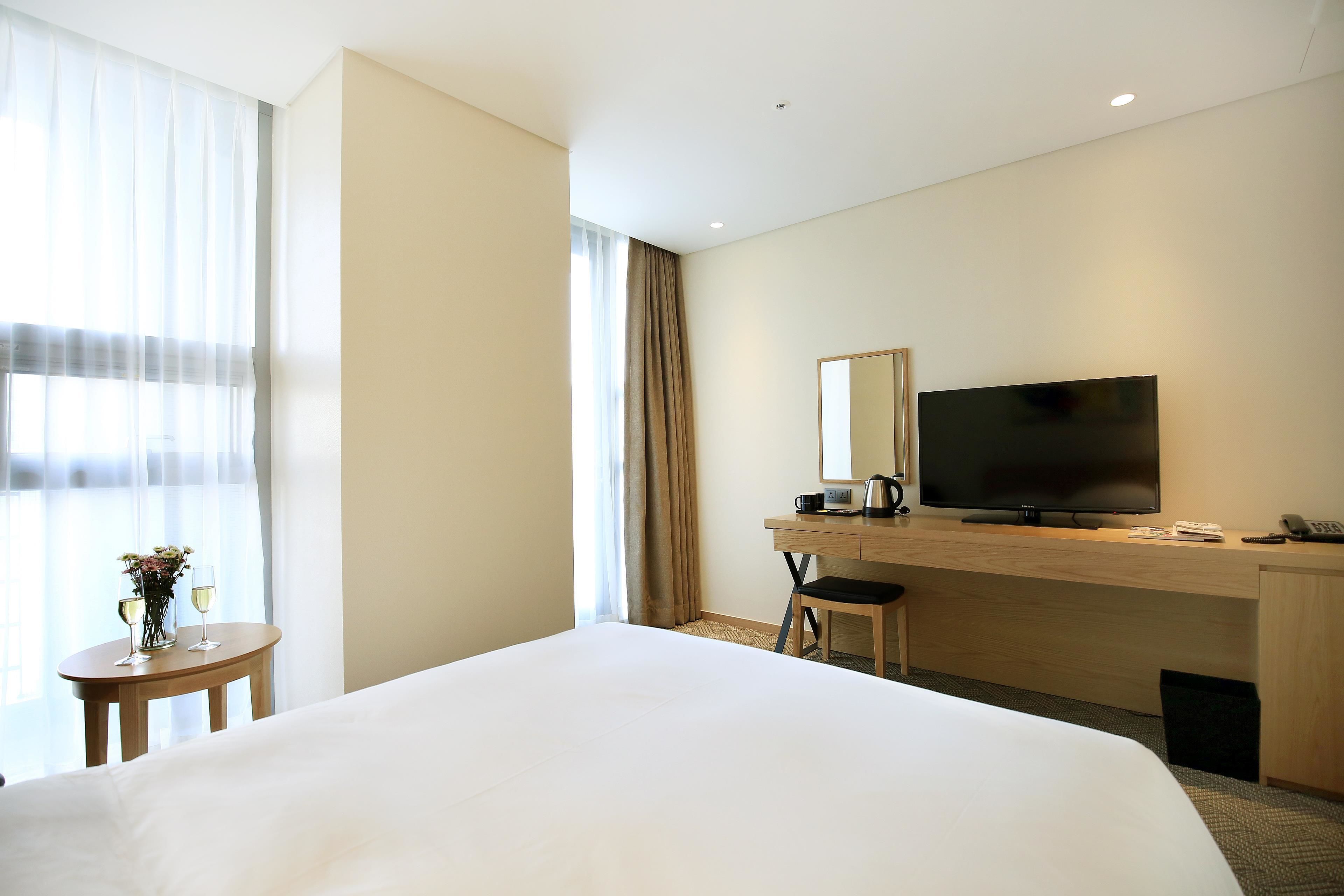 A hotel room with a large bed, a desk with a television, a small table with a vase and glasses, and a window with curtains.