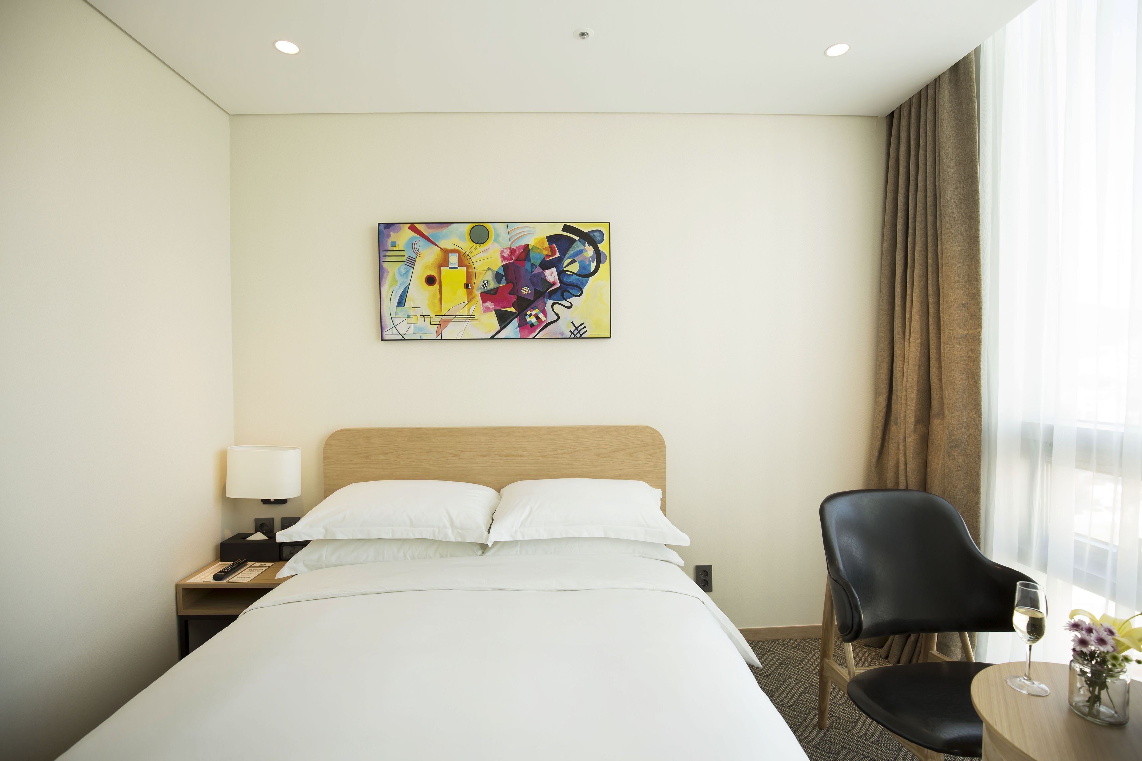 Quarto de hotel com cama, uma cadeira, mesa e uma taça de vinho, além de um quadro na parede.