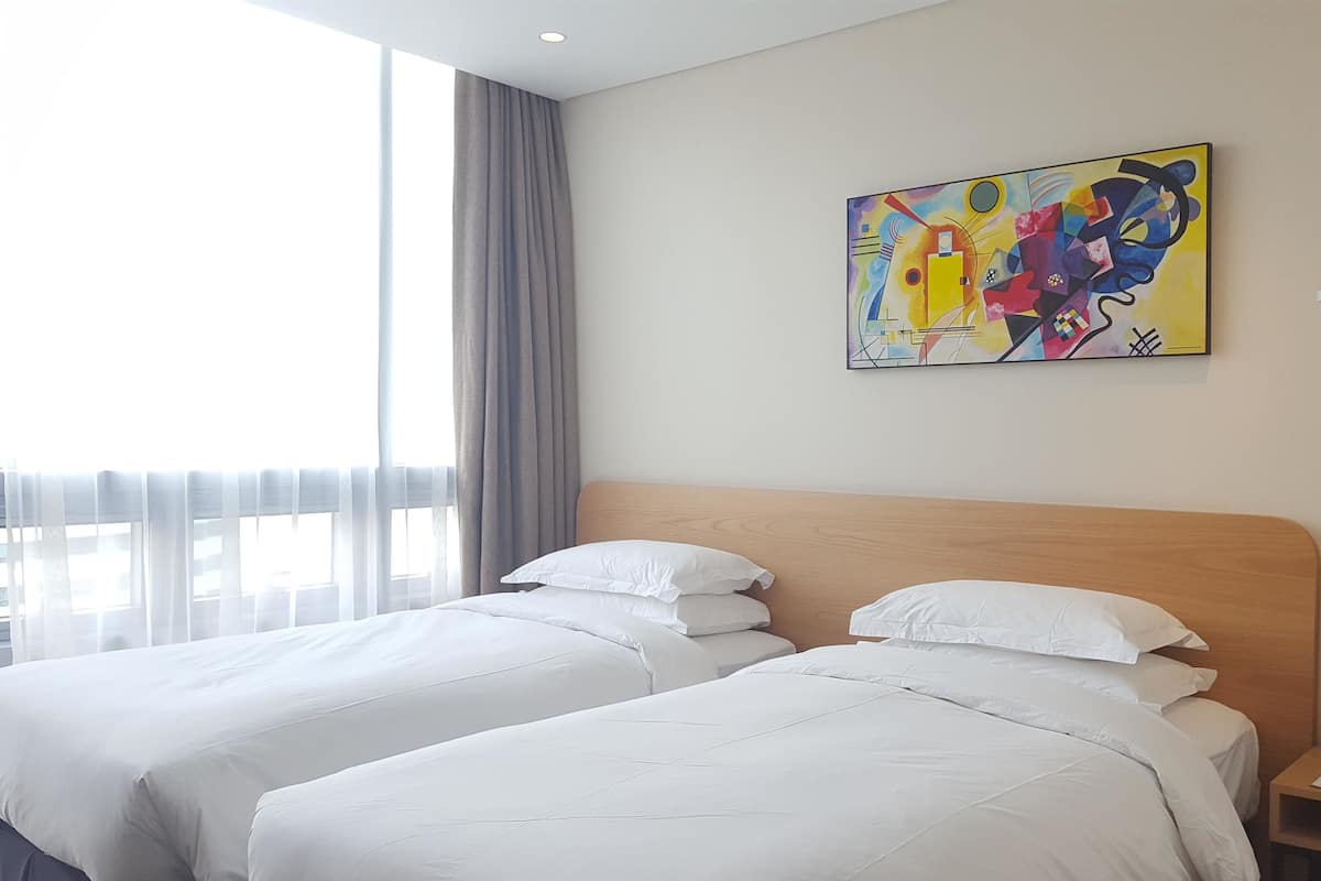 A hotel room with two beds, a large window with sheer curtains, and a colorful abstract painting on the wall.