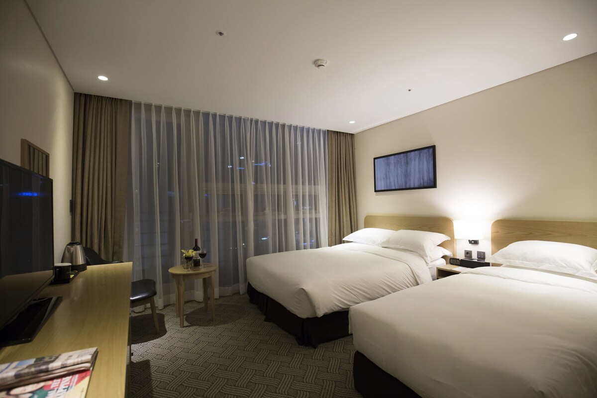 Hotelkamer met twee bedden, een bureau, een televisie, een klein tafeltje met een lamp en een raam met gordijnen.