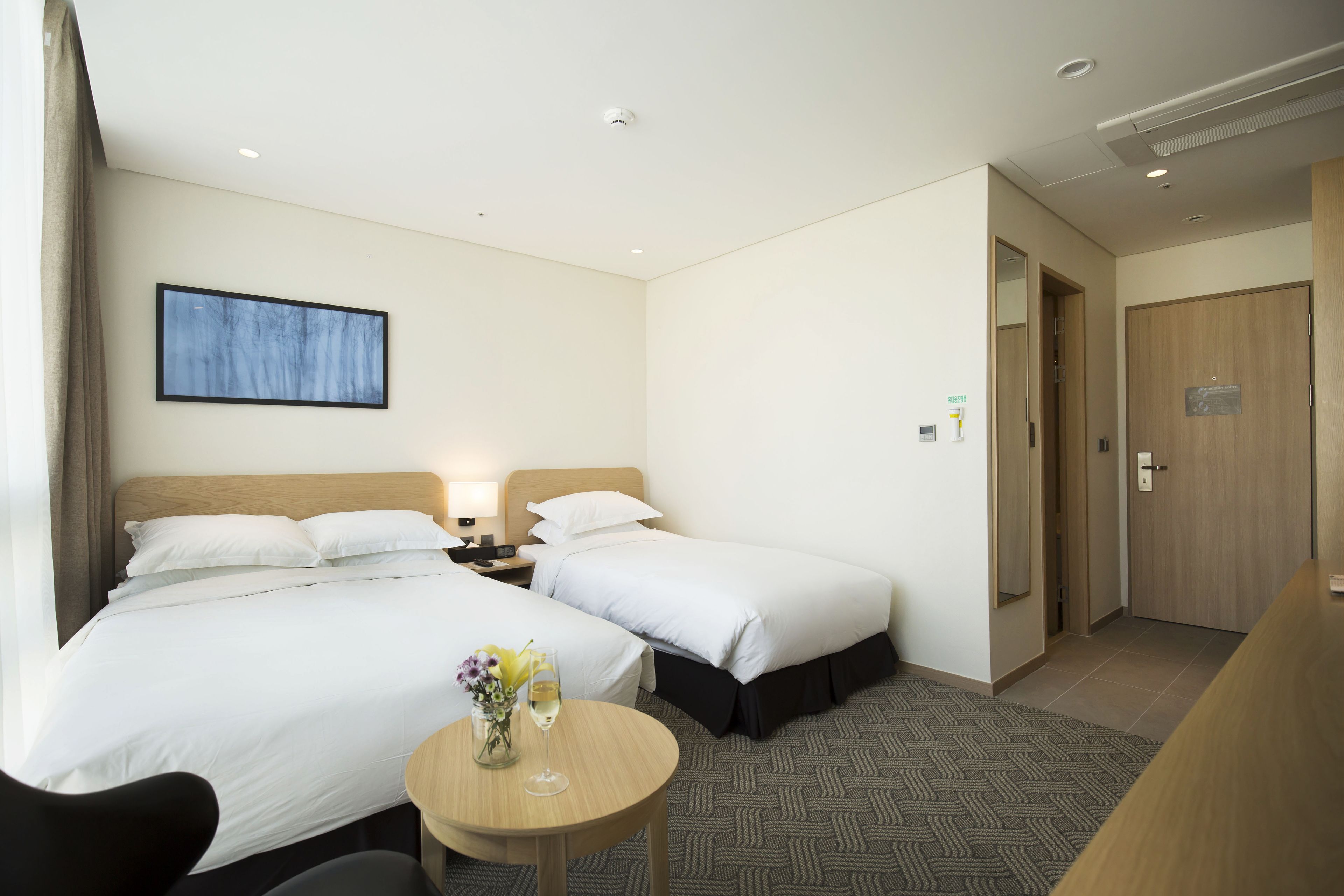 A hotel room with two beds, a small table with a vase and a glass, and a wooden door.