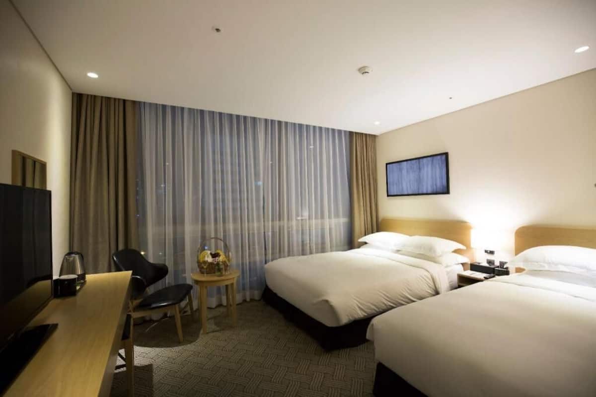 A hotel room with two beds, a desk, a chair, a small table with a lamp, and a TV mounted on the wall.